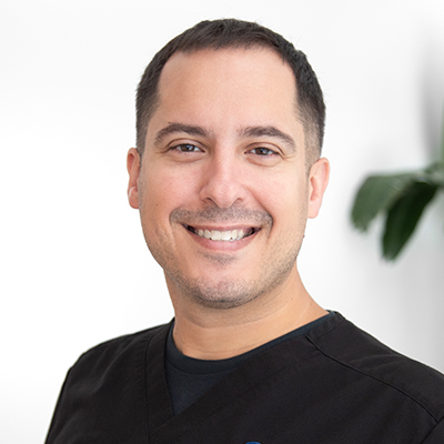 Frank Lombardo, OTR/L, an Occupational Therapist wearing a black scrub top embroidered with the "Prestige Health & Wellness" logo and his name, title, and specialization. He is smiling, looking directly at the camera, and standing in front of a white background with a green plant blurred in the distance on the right side