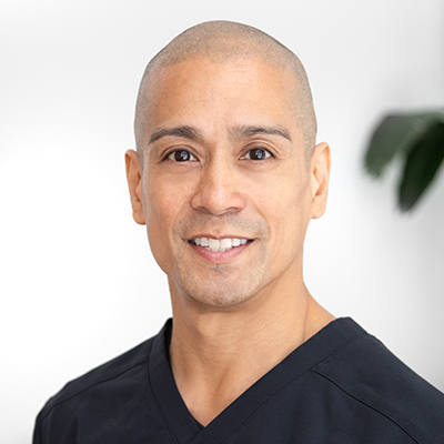 This image shows Dr. Rey Gayo, DC, a chiropractor, wearing a black scrub top. He is smiling, looking directly at the camera, and standing against a white background with a blurred green plant in the background on the right side.