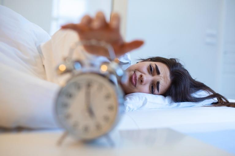 Woman in bed hitting alarm clock with a look of dread, representing sleep problems and insomnia in need of treatment.